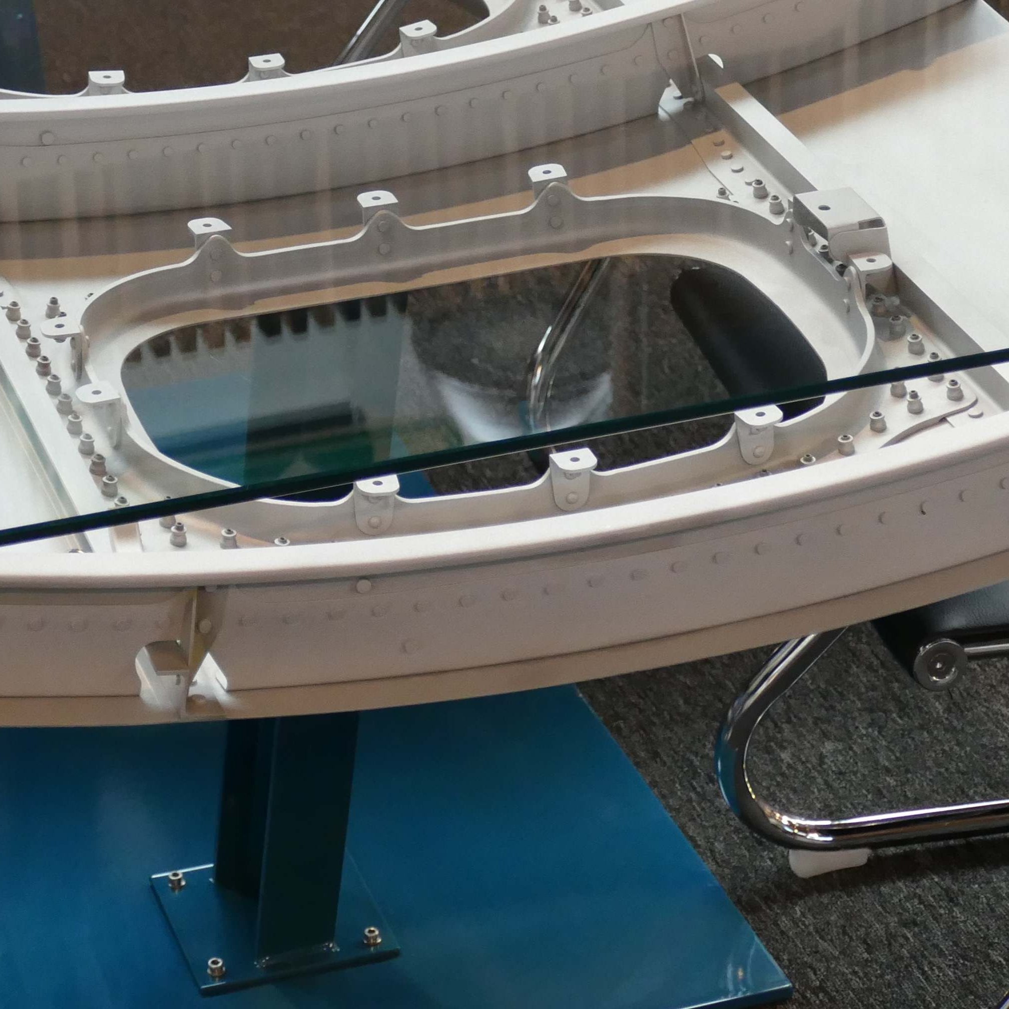 Designer-Bürotisch mit weißem Metallrahmen und Glasplatte, blauem Standfuß, modernem Bürostuhl auf grauem Teppich.