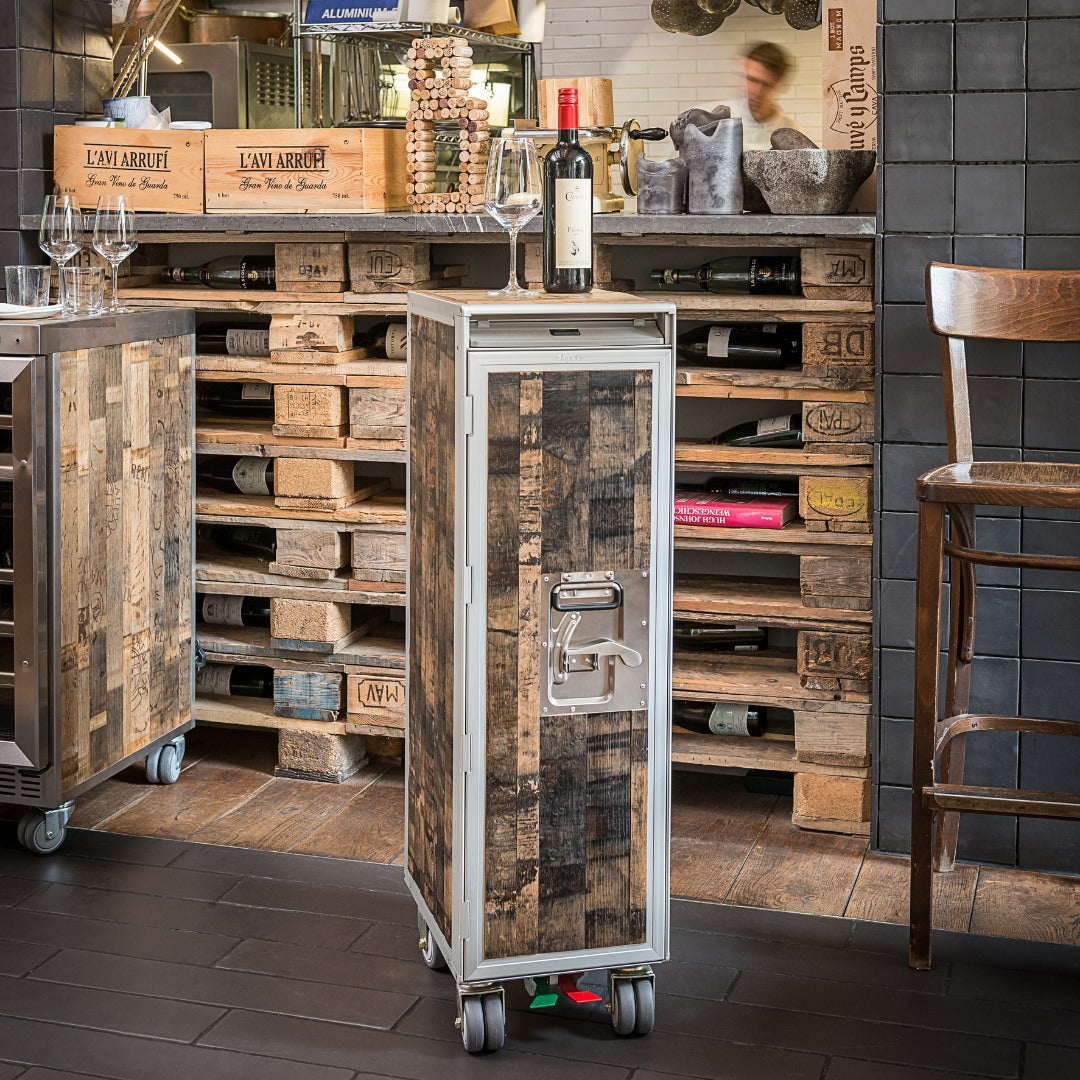 Eleganter Holztrolley mit Weinflasche und Glas vor rustikalem Regal mit Weinkisten und Flaschen