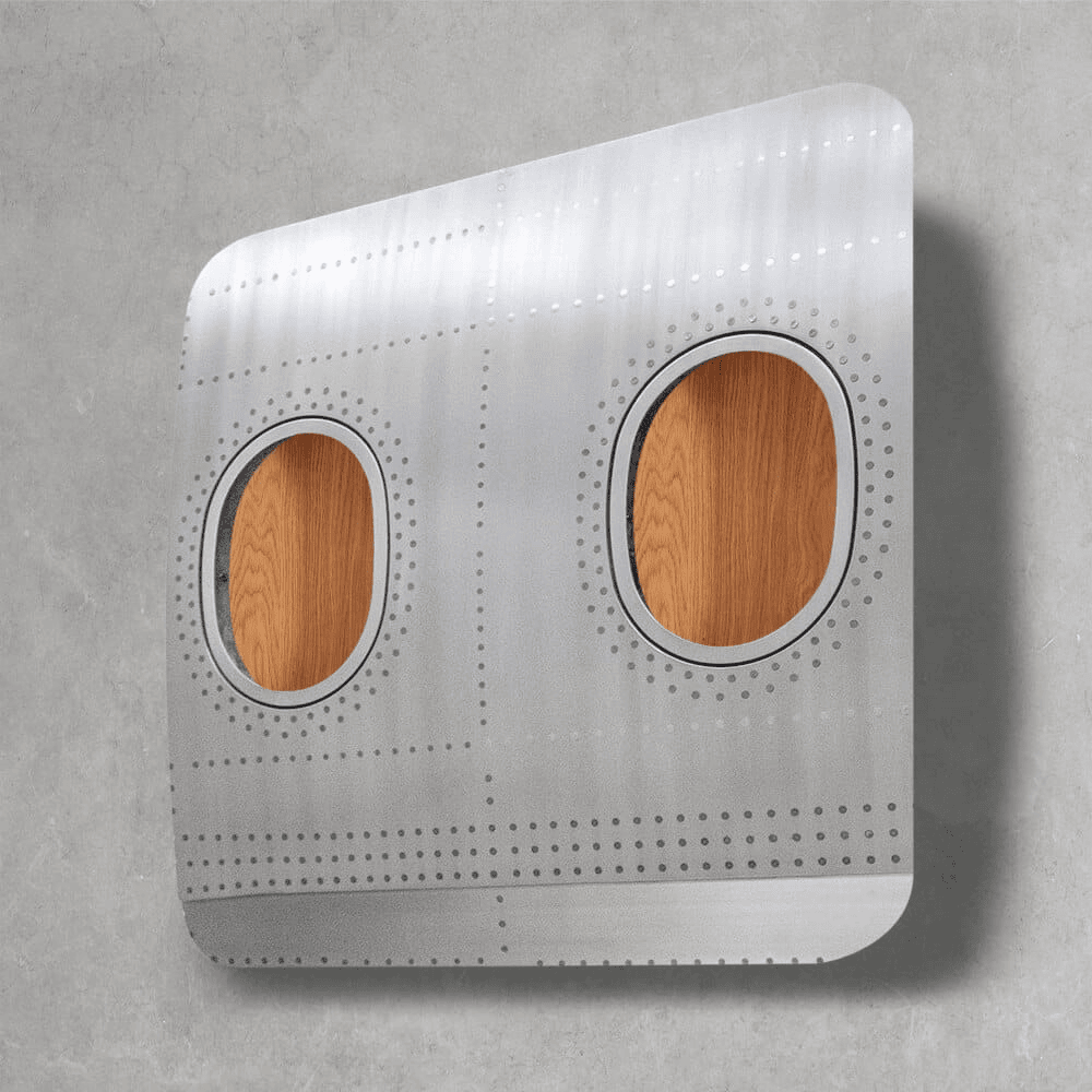 Rechteckiges Flugzeugrumpfsegment aus Metall mit Nieten und zwei ovalen Fenstern vor grauer Wand