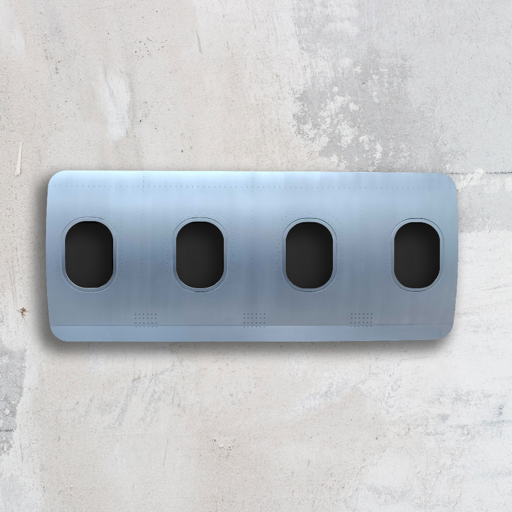 Horizontale Flugzeugrumpfsektion in Hellblau-Grau mit vier ovalen Fenstern vor einer Betonwand.