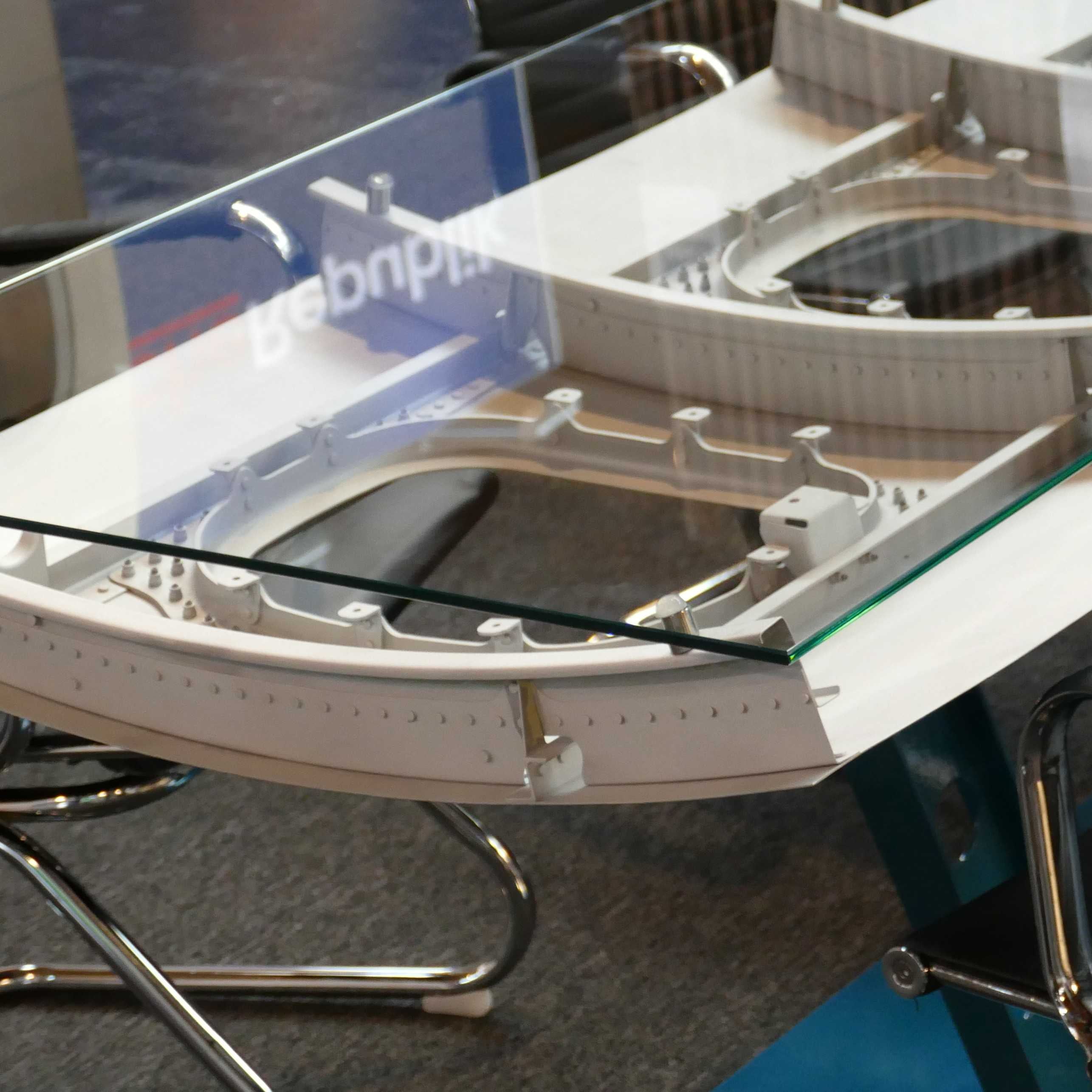 Glasplatte mit weißem Metallrahmen und schwarzen Bürostühlen auf grauem Teppich mit blauer Fläche.