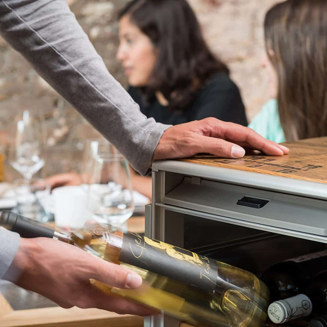 Hand hält eine Flasche Chamois Terre Weißwein vor einem Weinkühlschrank, im Hintergrund unscharf Menschen mit Weingläsern.