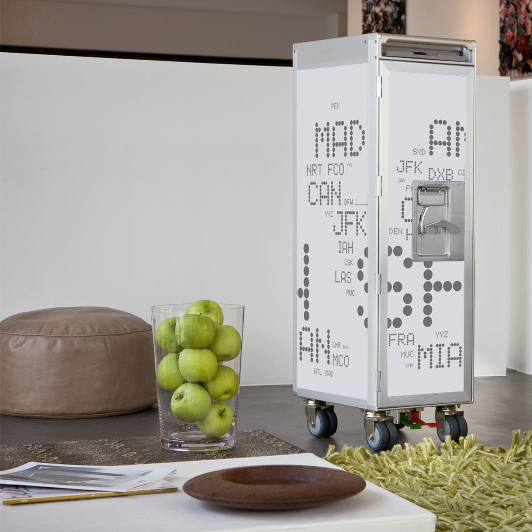 Moderner Wohnraum mit Airline-Trolley und Glasvase mit grünen Äpfeln auf weißem Tisch, Pouf und Teppich.