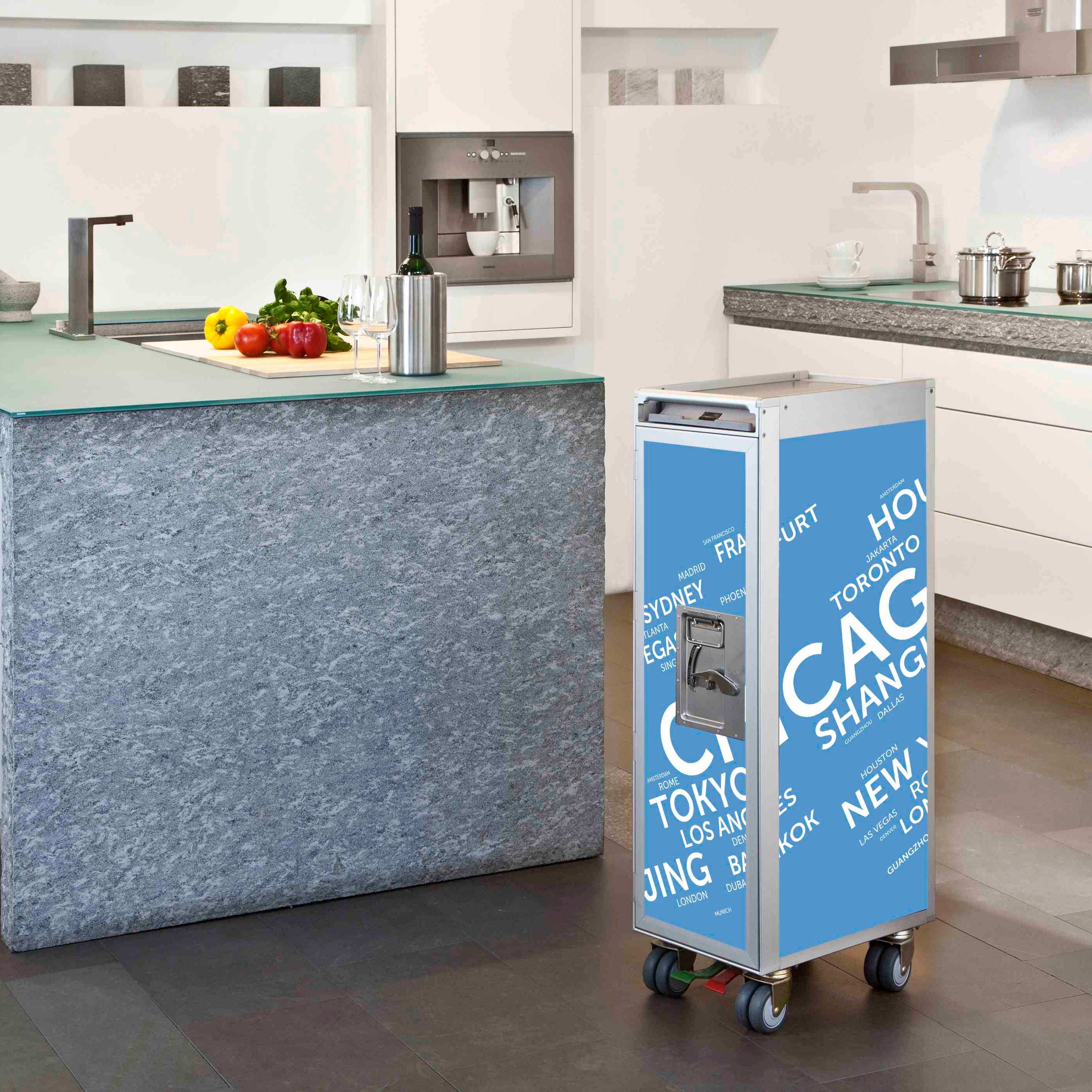 Ein moderner Küchenraum mit blauem Airline-Trolley, bedruckt mit Städtenamen, neben einer Kücheninsel mit Gemüse und Wein.