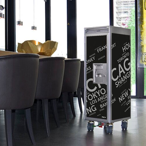 Moderner Innenraum mit schwarzem Rollwagen aus Flugzeugteilen, bedruckt mit Städtenamen, neben Polsterstühlen vor Fenstern.