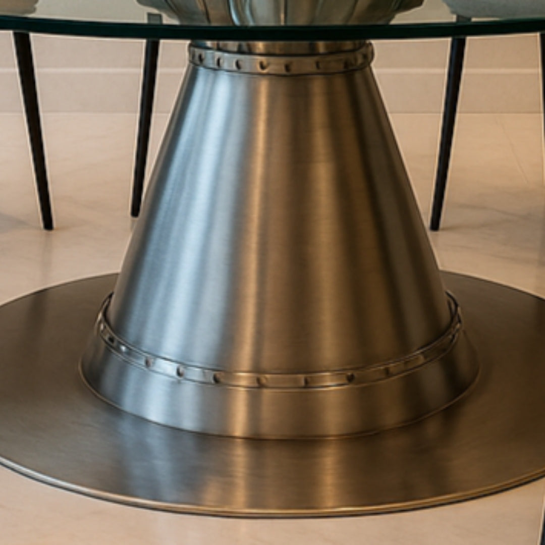 Moderner Tisch mit konischem Metallfuß aus gebürstetem Aluminium und Glasplatte auf heller Marmorfliese.