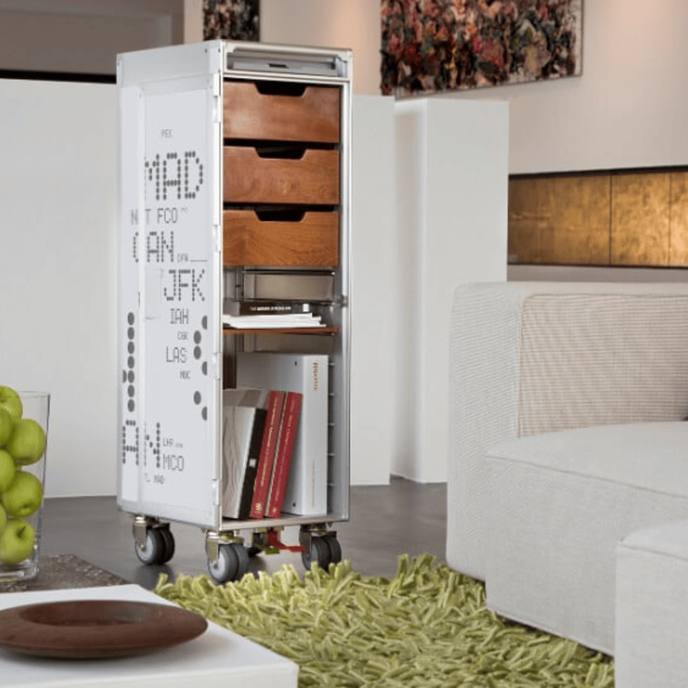 Moderner Wohnraum mit weißem Skypak Airline Trolley aus Metall und Holz, dekoriert mit Flughafen-Codes, neben beigem Sofa mit gestreiftem Kissen auf grünem Teppich.