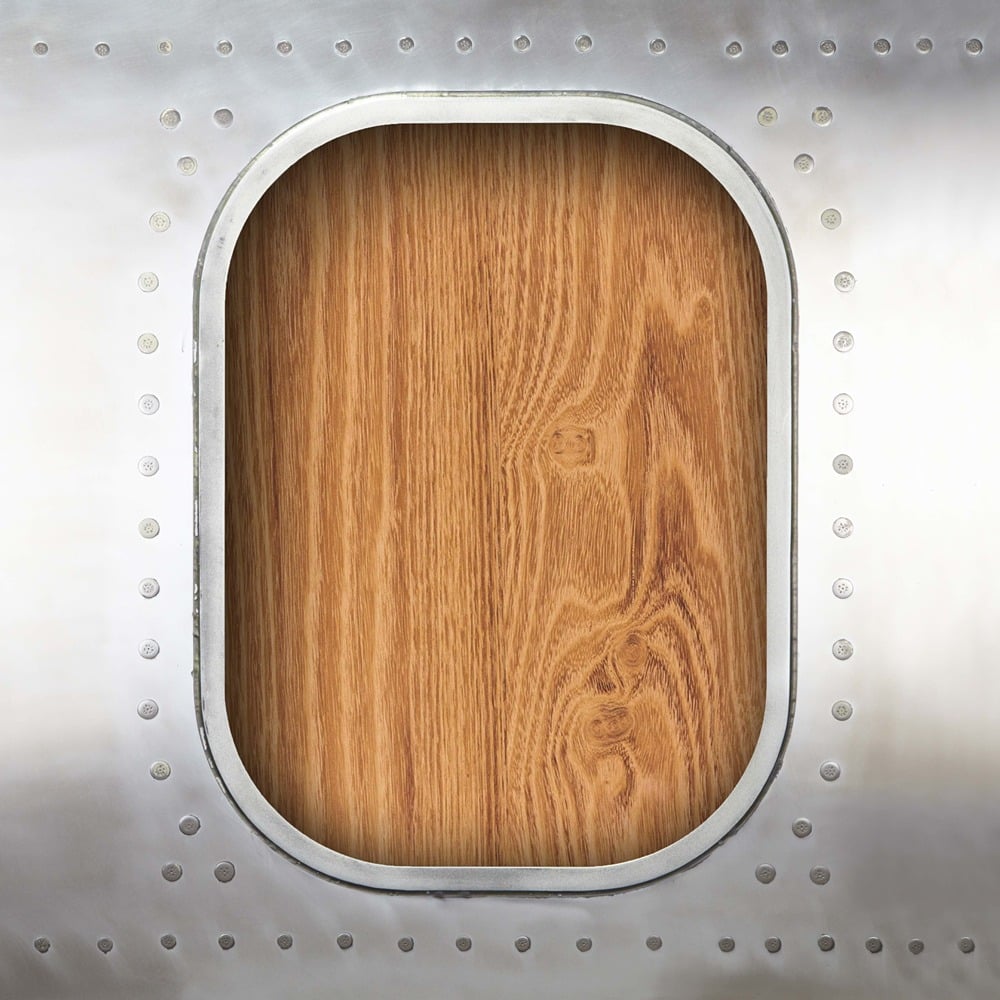 Nahaufnahme eines rechteckigen Flugzeugfensters mit abgerundeten Ecken und silbernem Metallrahmen mit Nieten, davor eine Holztafel in warmen Brauntönen.