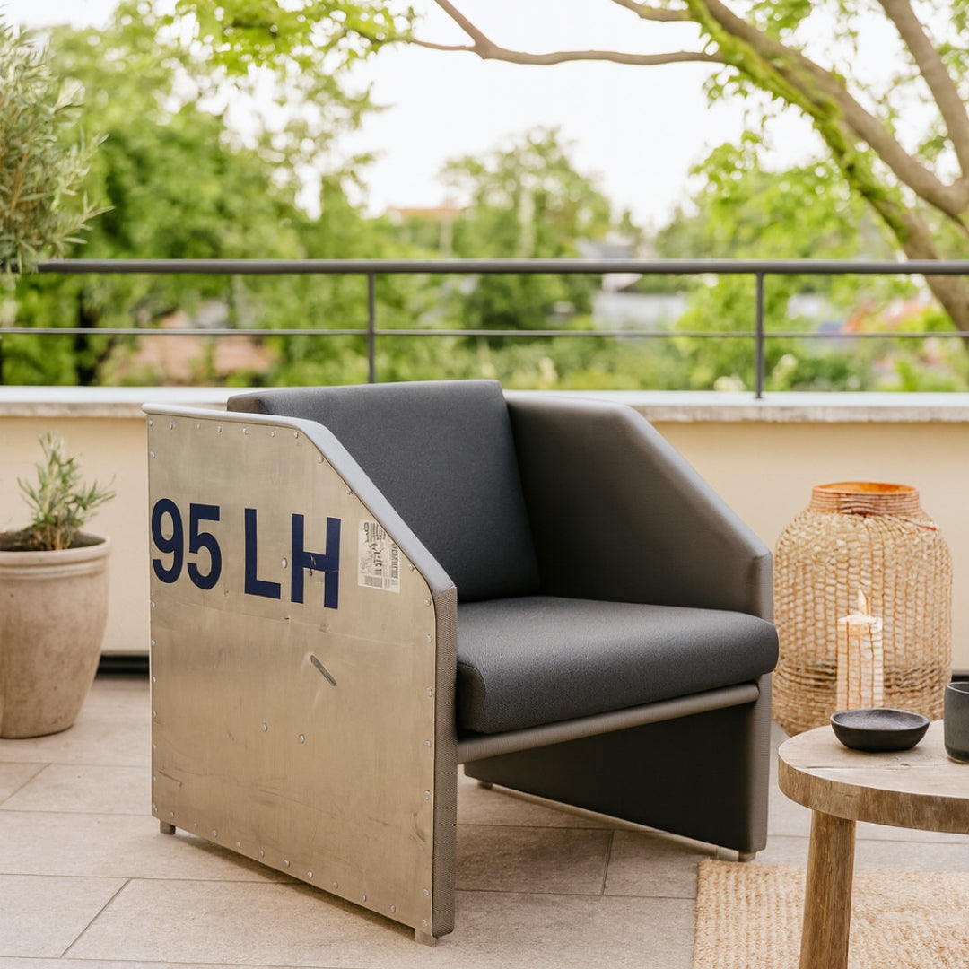 Moderner Outdoor-Armlehnstuhl mit dunkelgrauer Polsterung, metallischen Seiten mit blauer Beschriftung 95 LH, rundem Holztisch mit schwarzer Schale und Tasse, großer geflochtener Laterne mit Kerze und grüner Topfpflanze auf einer Terrasse mit Kacheln und schwarzem Geländer.