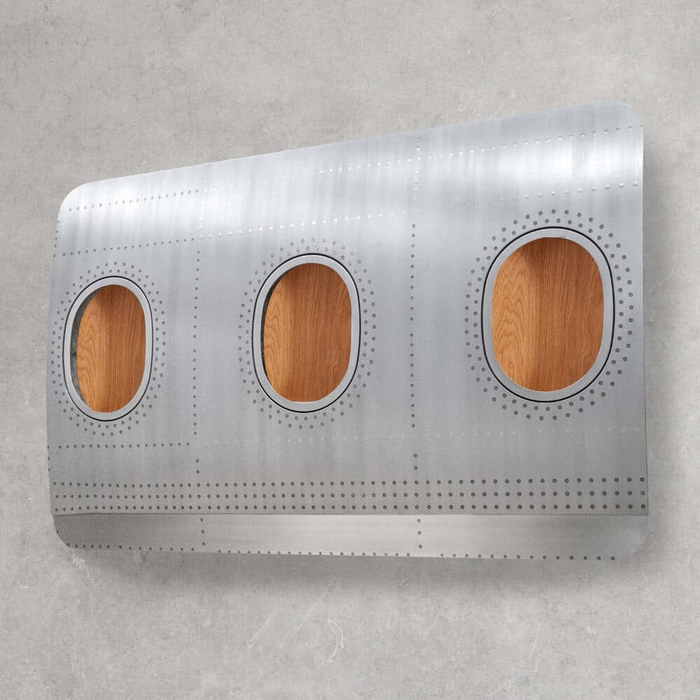 Segment eines silbernen Flugzeugrumpfs mit drei ovalen Fenstern mit Holzeinsätzen vor einer hellgrauen Wand