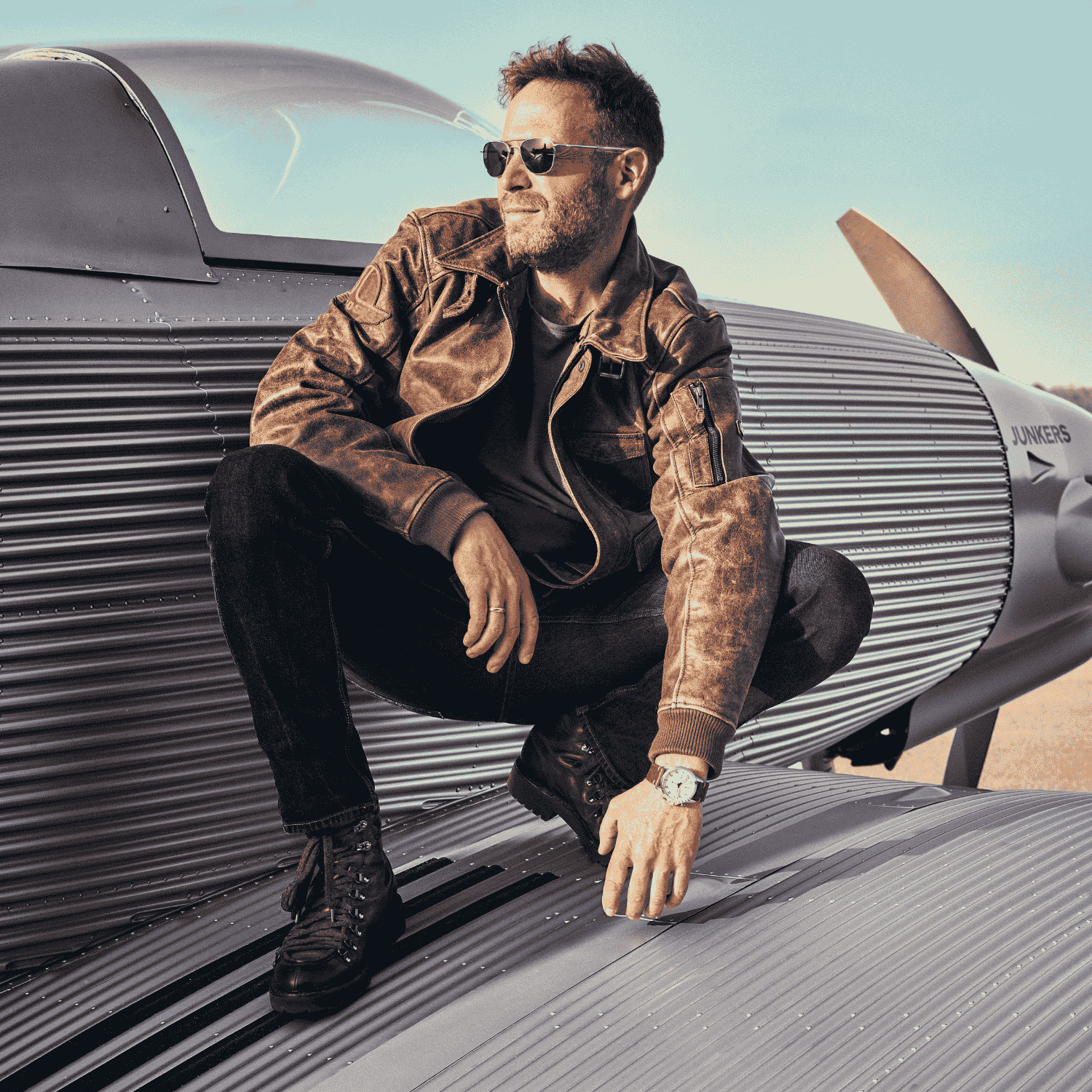 Mann in brauner Lederjacke und Sonnenbrille sitzt auf dem Flügel eines silbernen Vintage-Flugzeugs mit JUNKERS-Schriftzug.