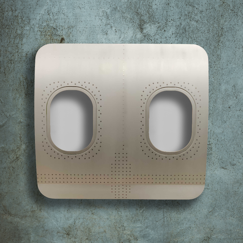 Metallsegment eines Flugzeugrumpfs mit zwei ovalen Fenstern und Nieten vor blauer Betonwand.