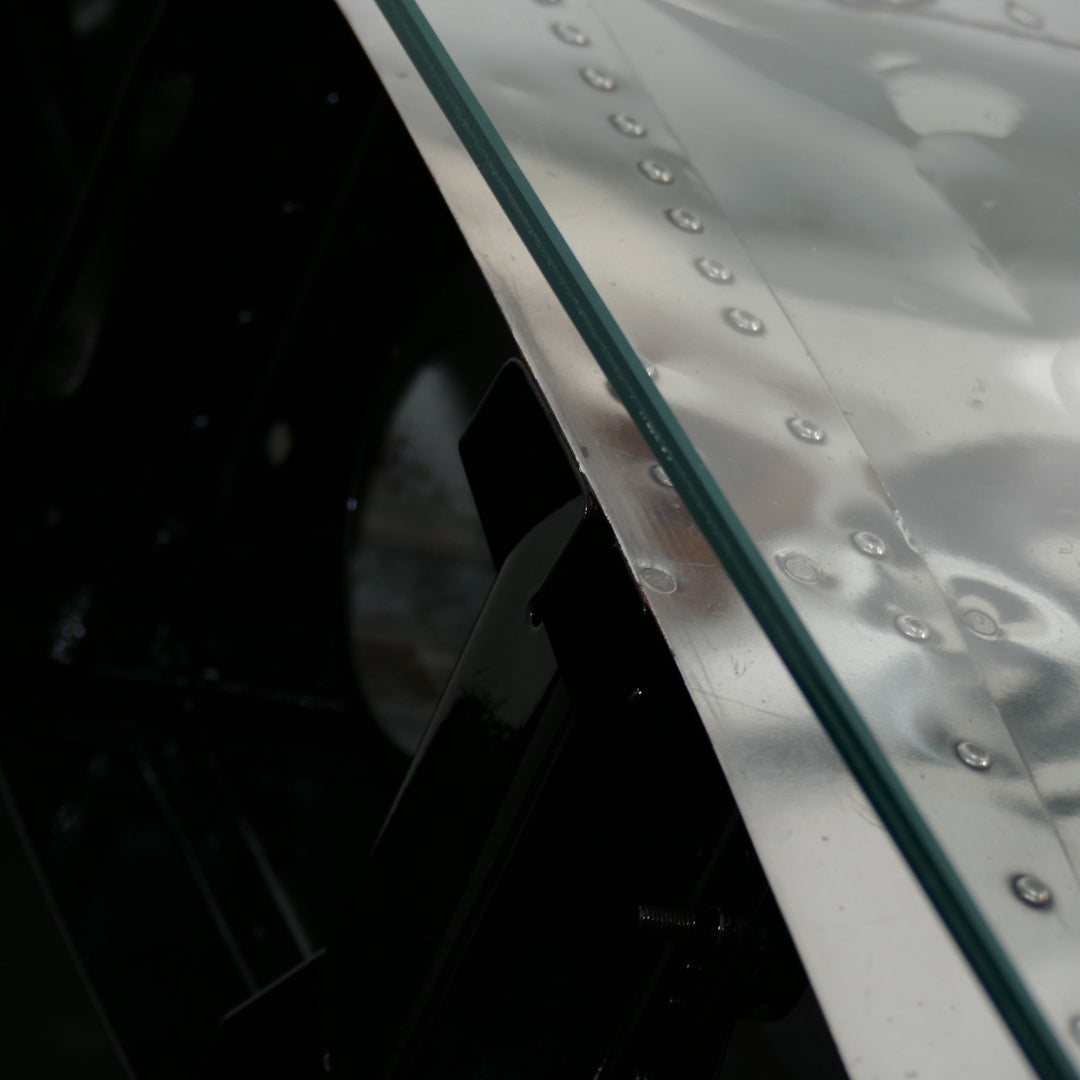 Nahaufnahme einer glänzenden, genieteten Metalloberfläche mit Glasplatte und schwarzer Metallstütze im technischen Design.