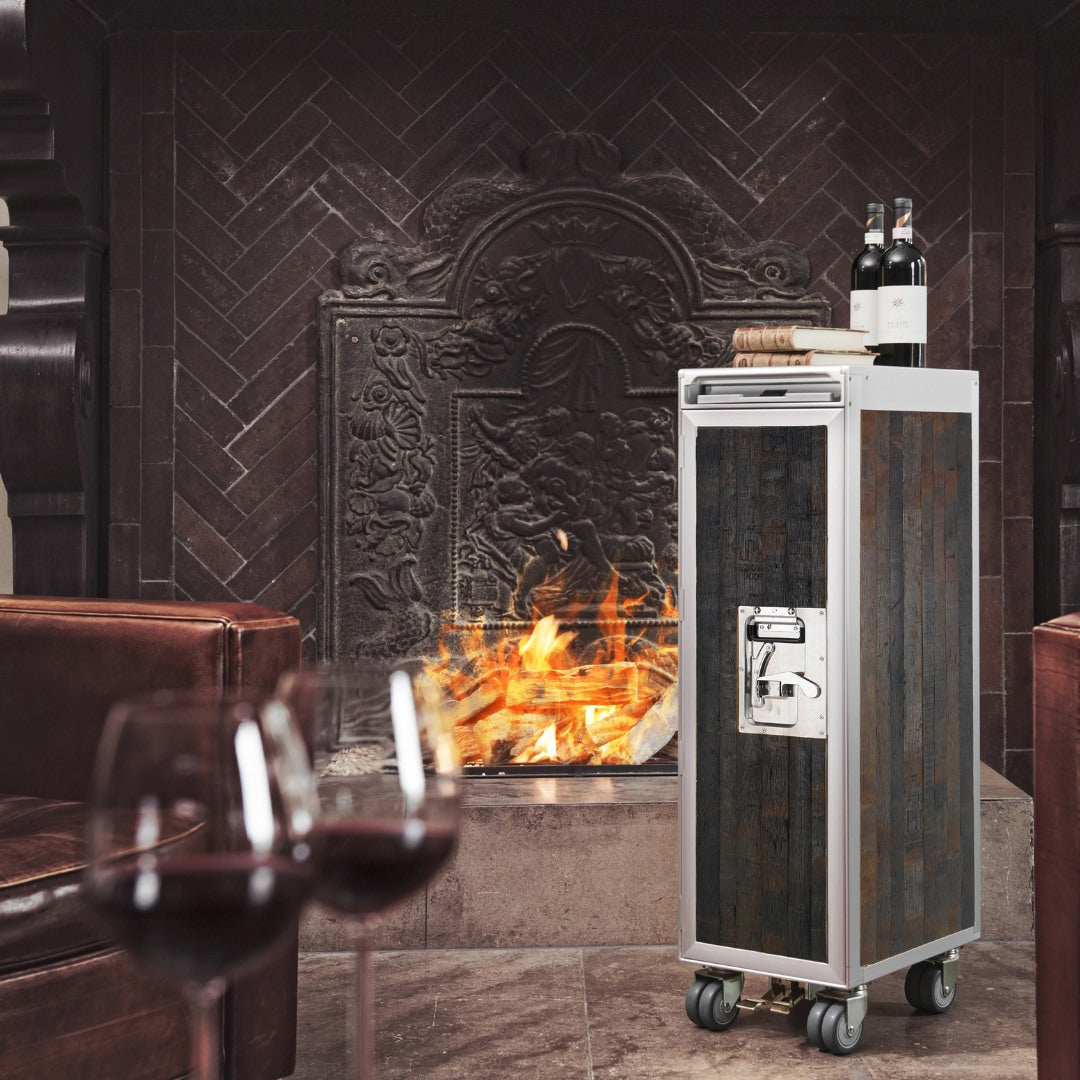 Eleganter Weinschrank aus Holz und Metall auf Rollen vor einem Kamin mit brennendem Feuer.