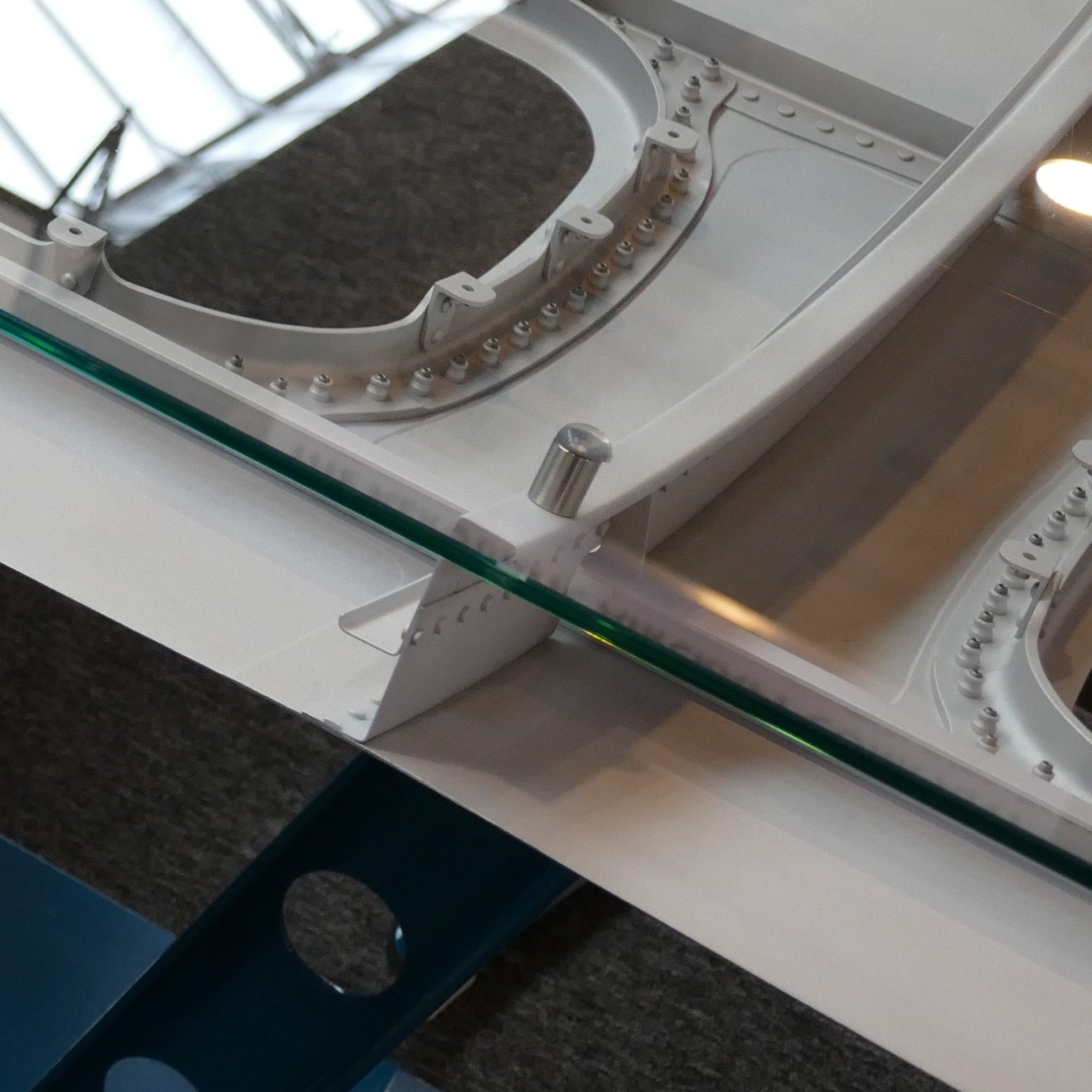 Nahaufnahme einer weißen, gebogenen Metallstruktur mit zahlreichen Nieten auf blauem Ständer hinter Glasbarriere.